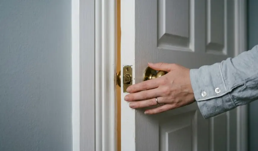 The Quiet Click – Ending the Day That Belongs to Others A woman's hand resting on a closed bedroom door, wearing a soft pajama sleeve with mother-of-pearl buttons, blurred bedroom behind.